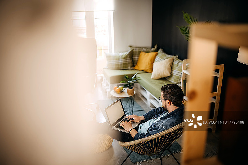 戴眼镜的年轻络腮胡男子坐在家中的扶手椅上使用笔记本电脑图片素材