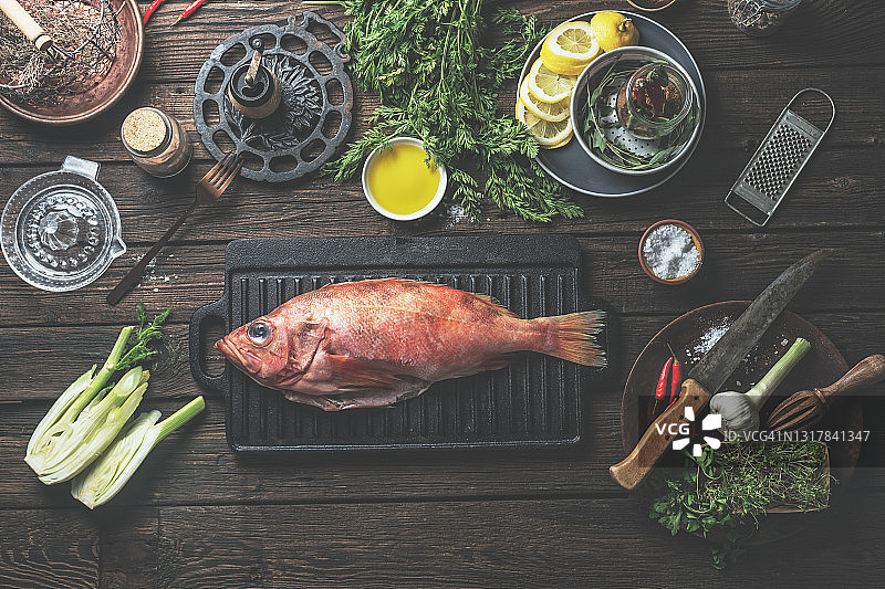 乡村木制厨房桌上的黑色铸铁烤架上的完整生红鱼，配有食材和复古厨具图片素材