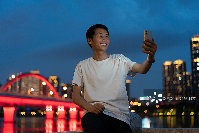 坐在栏杆上用智能手机进行视频通话的年轻亚洲男人图片素材