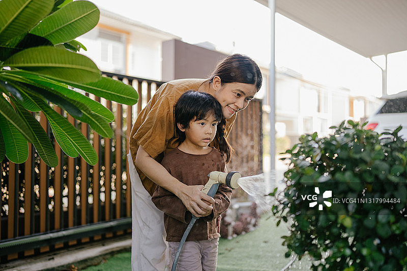 男孩和他的母亲在阳光明媚的后院用花园软管浇灌植物图片素材