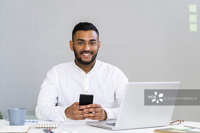 年轻男商人坐在办公室桌旁，用智能手机操作笔记本电脑图片素材