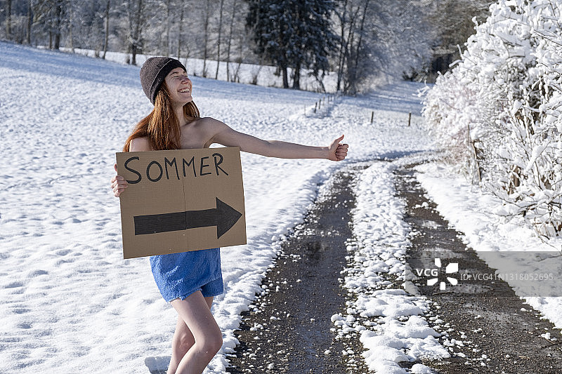 在白雪覆盖的道路上搭便车的微笑女人，手里拿着标牌图片素材