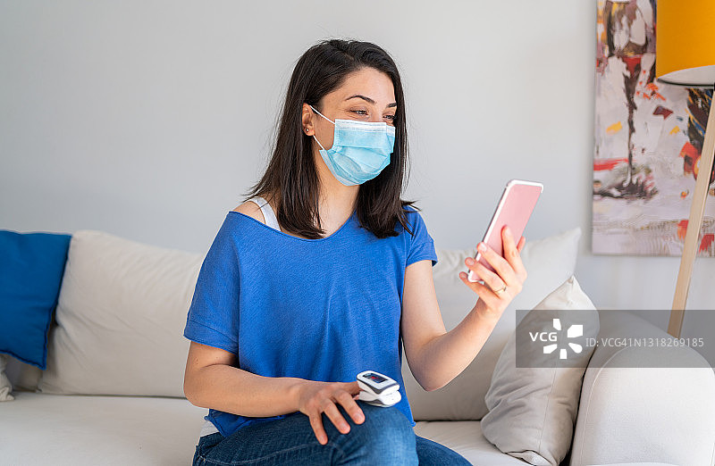 戴防护口罩的女人在家用数字脉搏血氧仪测量、监测氧饱和度。患有冠状病毒、新冠疫情隔离的女人正在使用智能手机进行远程医疗视频通话，咨询医生图片素材