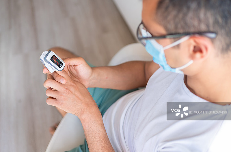 戴防护口罩的男人在家中使用数字脉搏血氧仪测量、监测氧饱和度。患有冠状病毒的男人，新冠疫情隔离封锁。在家中使用数字脉搏血氧仪测试血液中的氧气含量和脉搏频率。图片素材