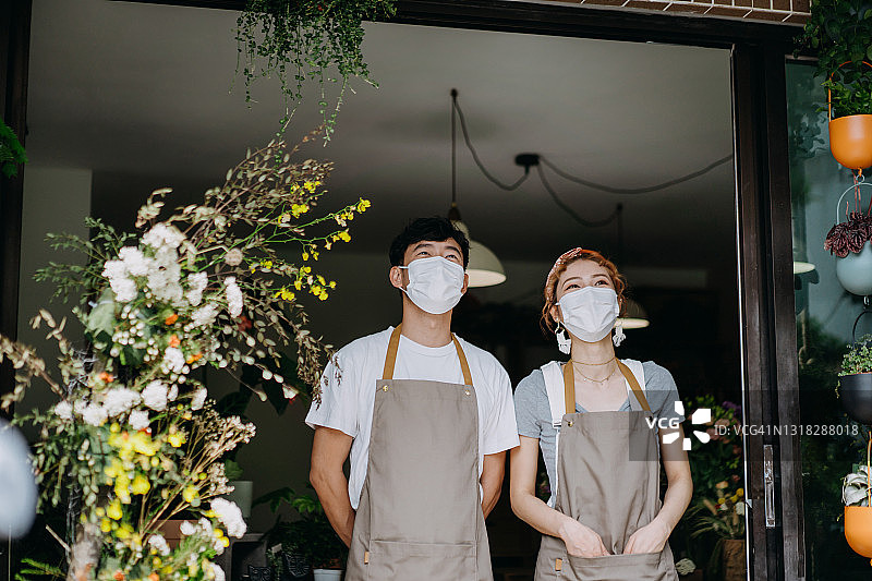 自信的亚洲年轻夫妇经营一家小型花店。戴着防护口罩以预防病毒。站在店前，面带微笑和积极的情绪看着远方。合作与团队合作。新冠疫情后重新开业概念图片素材