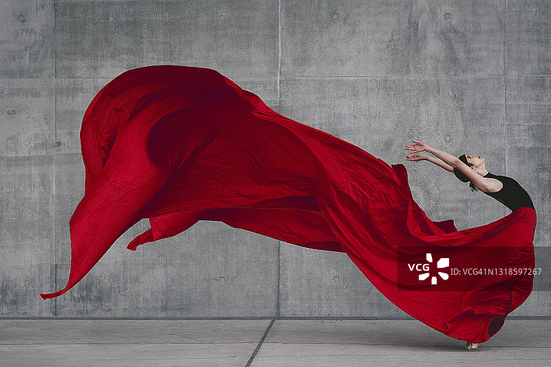 在灰色水泥墙前跳舞的女芭蕾舞演员，脚尖有红色面纱图片素材
