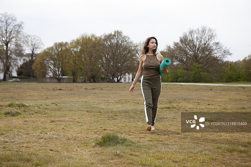 一位年轻女子赤脚走在公园里，胳膊下夹着卷起来的瑜伽垫图片素材