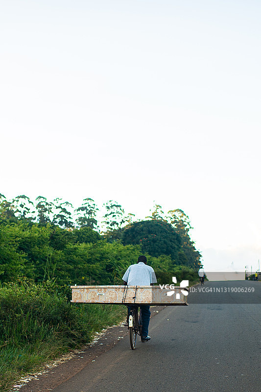 男子骑自行车在沥青路边行驶，车后驮着棺材图片素材