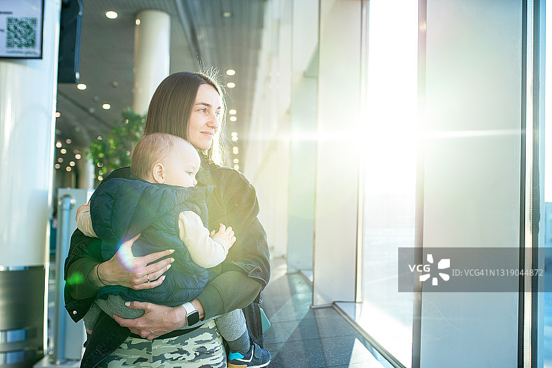 日出时，抱着婴儿站在机场航站楼窗户旁的女人图片素材