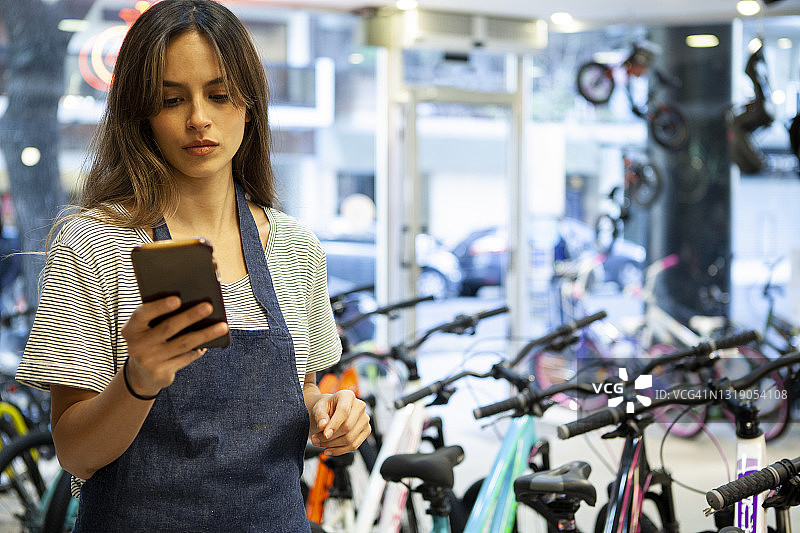 专注的女性店主在自行车商店里走动时查看手机上的内容图片素材