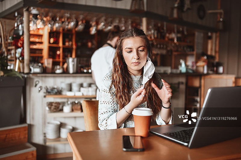 在咖啡吧里，女人摘下口罩，一边用笔记本电脑一边喝咖啡图片素材