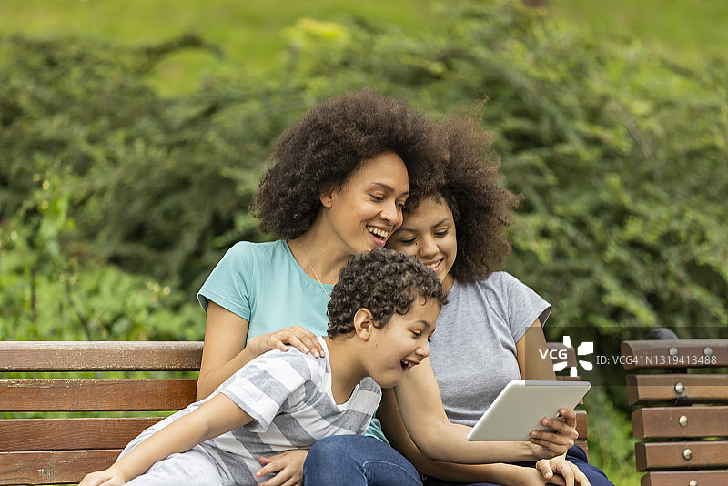 非洲裔美国妇女与儿子和女儿坐在公园里使用平板电脑图片素材