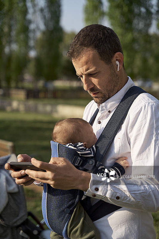 戴着入耳式耳机和男婴在公园使用手机的男人图片素材