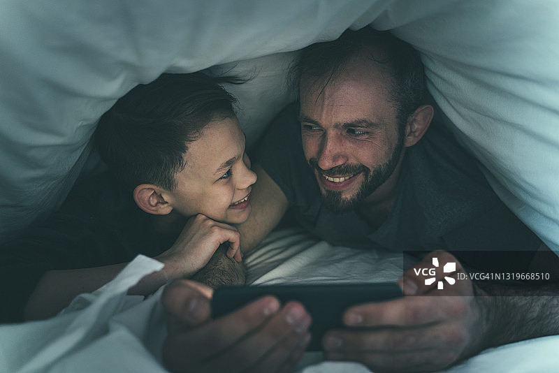 父子俩在家中在毯子下使用智能手机时互相看着对方图片素材