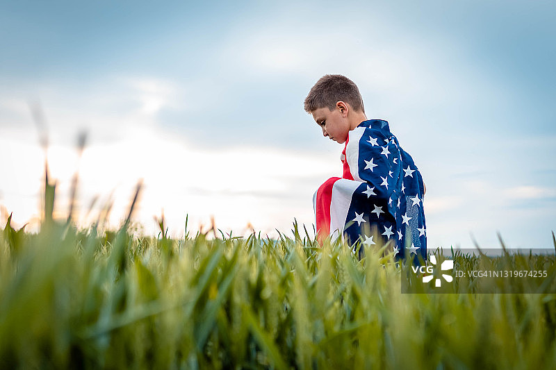 裹着美国国旗的悲伤男孩。他跪在田野上，看起来很悲伤图片素材