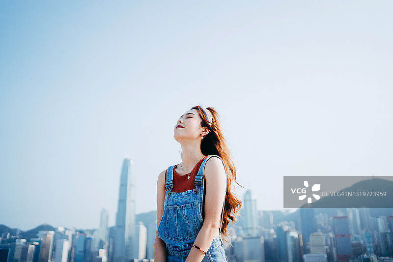 一位美丽的微笑亚洲年轻女性闭着眼睛，深吸一口气，享受着新鲜的空气和柔和的微风，背景是壮观的维多利亚港海滨城市天际线图片素材
