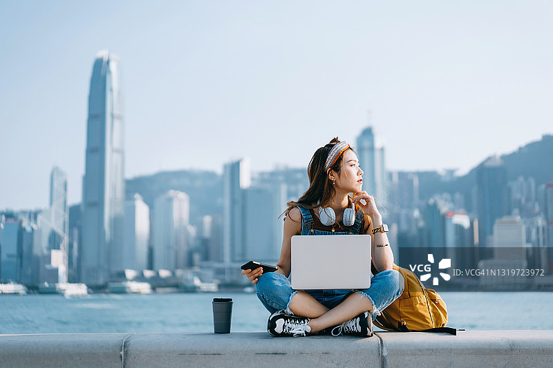 年轻漂亮的亚洲女子在长廊边盘腿而坐，以城市天际线为背景。她戴着耳机，使用智能手机和笔记本电脑工作，旁边放着一杯咖啡。若有所思地看向远方。生活方式与科技图片素材