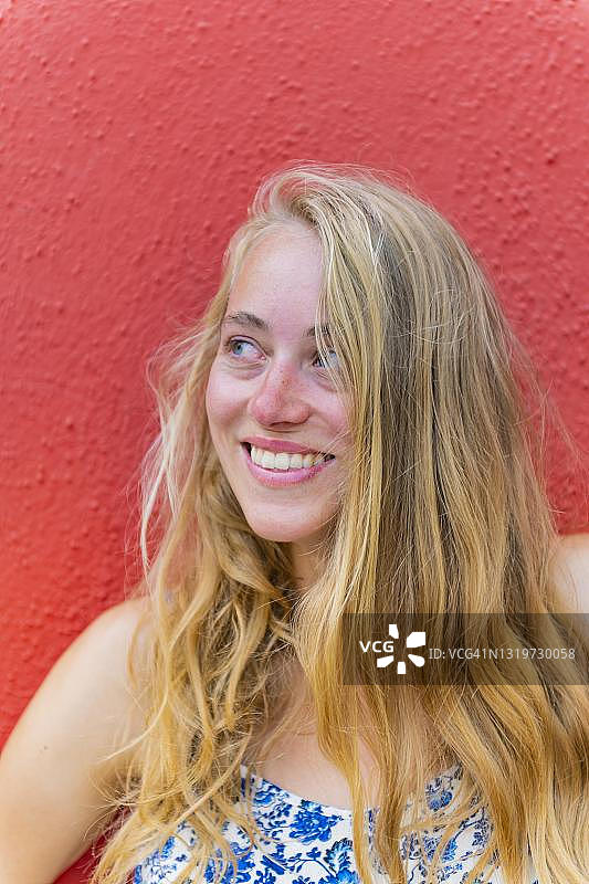 在红色墙壁前微笑的金发年轻女子，意大利威尼托布拉诺岛图片素材