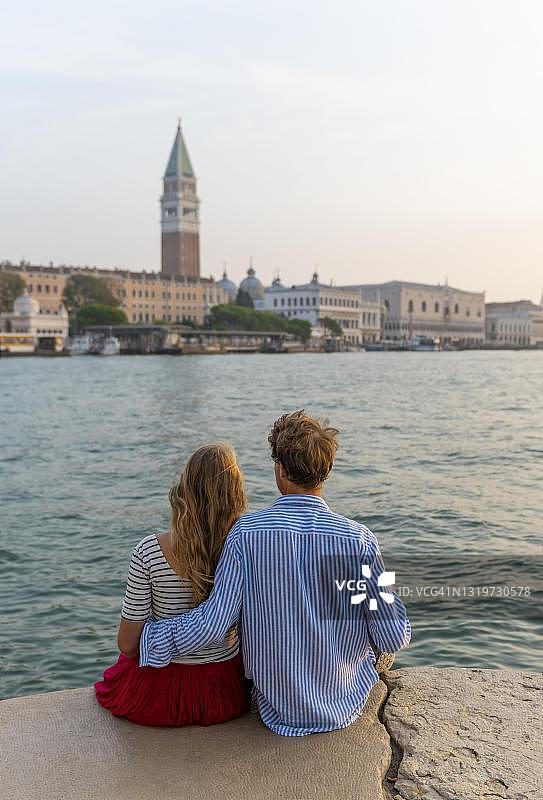 年轻夫妇坐在海边欣赏圣马可广场和钟楼的景色，意大利威尼托图片素材