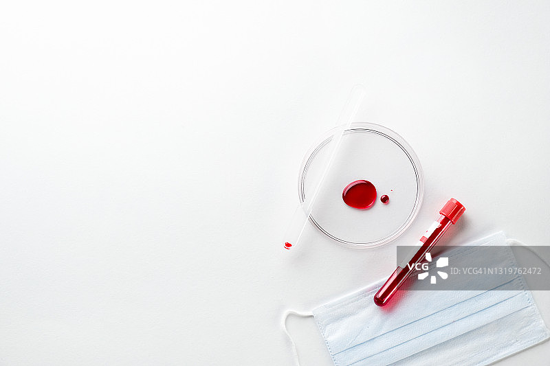 白色背景下玻璃培养皿中的一滴血。装有医疗样本的试管用于检查。旁边是一个医用防护一次性口罩。医疗保健概念。图片素材