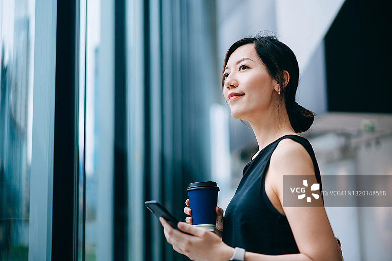 在现代办公室里拿着智能手机，自信且有抱负的亚洲女商人站在窗边图片素材