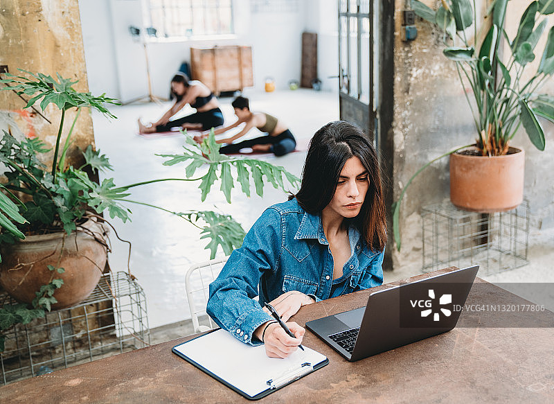 年轻女子在多功能空间使用笔记本电脑工作，其他人正在背景中训练图片素材