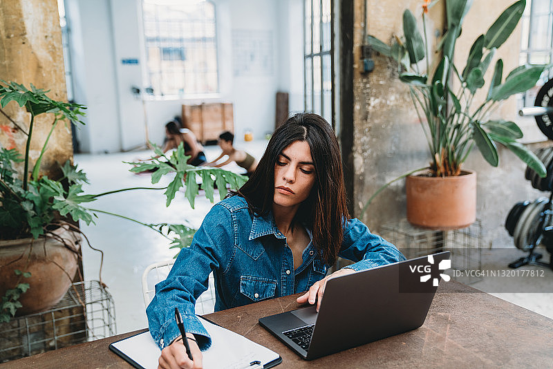 年轻女子在多功能空间使用笔记本电脑工作，其他人正在背景中训练图片素材