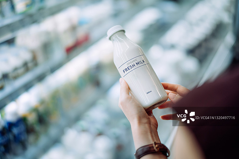 年轻亚洲女性在超市购物，肩上视角。手拿一瓶有机鲜牛奶，正在阅读营养标签。每日食品采购，健康饮食图片素材