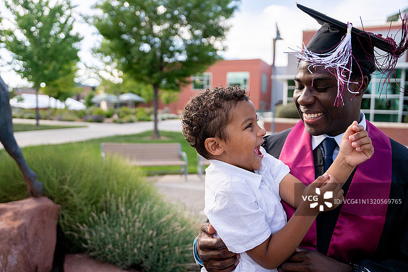 戴着毕业帽和长袍的非裔美国男大学生在毕业当天抱着他三岁的儿子玩耍图片素材