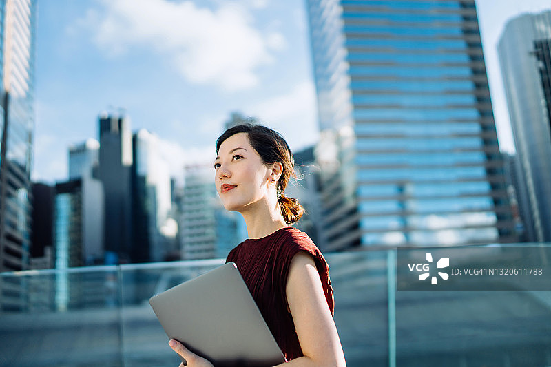 自信而现代的年轻亚洲女商人拿着笔记本电脑，在中央商务区通勤上班，站在城市的摩天大楼前。女性领导力，决心成功。移动办公图片素材