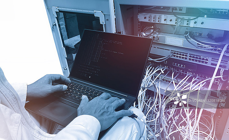 数据中心IT工程师使用笔记本电脑在服务器机架前进行日常维护检查和诊断，可见计算机硬件设备、宽带光缆和LED灯图片素材