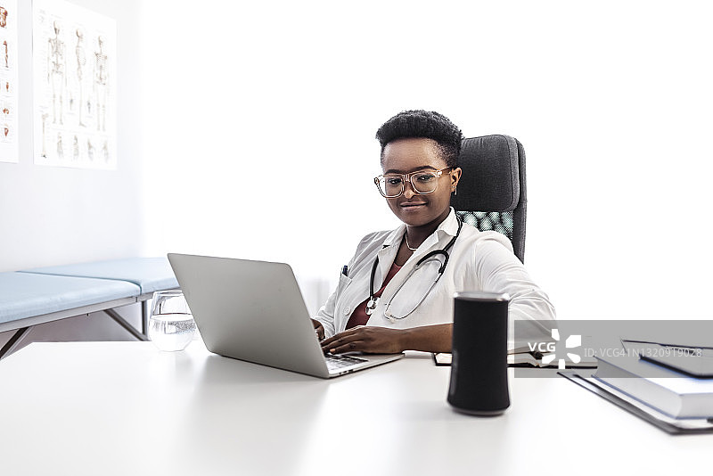 穿着白大褂，使用语音控制智能音箱的微笑黑人女医生图片素材