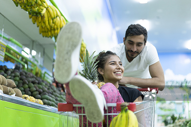 在杂货店购物的拉丁裔情侣，女子高兴地坐在购物车里图片素材