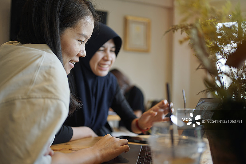 年轻的亚洲穆斯林自由职业女性在舒适的咖啡馆里用笔记本电脑工作和讨论图片素材