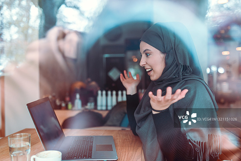 笑容满面的戴着头巾的穆斯林女性一边喝咖啡一边使用笔记本电脑图片素材