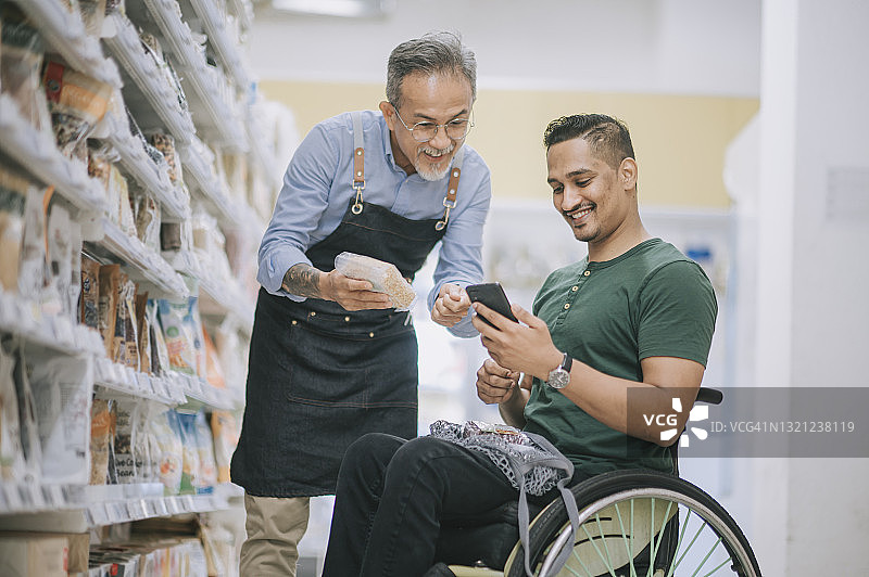 印度裔男子坐轮椅在超市用手机向年长男店员询问购物清单图片素材