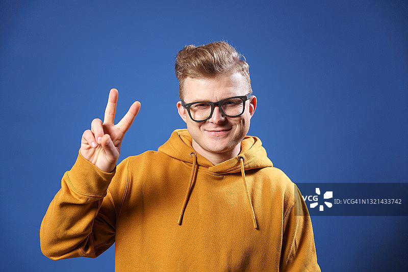 演播室蓝色背景中，戴眼镜、穿黄色连帽衫的男人用手指做出胜利手势图片素材