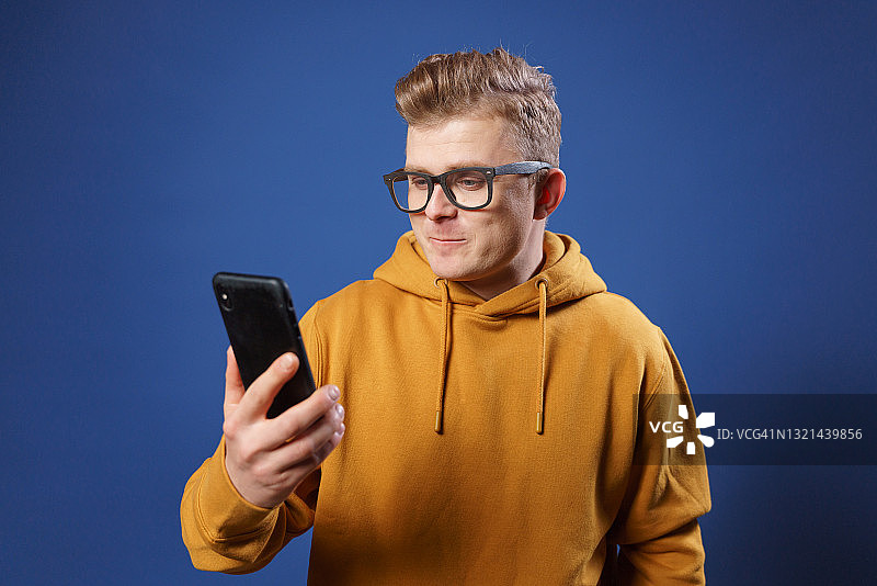 在蓝色背景工作室里看智能手机的戴眼镜黄帽衫男子图片素材