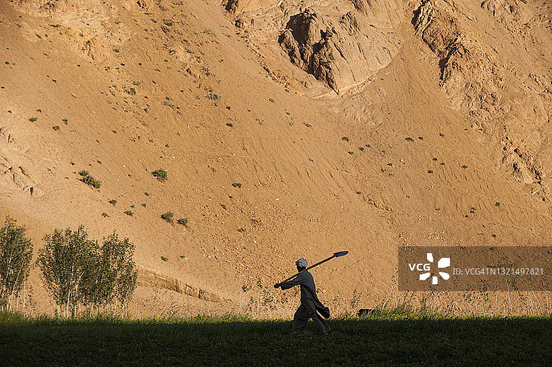 一名马铃薯农民在阿富汗巴米扬省干燥贫瘠的山坡上，扛着铁锹走过田野图片素材