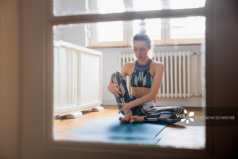 透过玻璃门看居家锻炼的女人：以束角式坐姿向上抬起右腿，疫情期间居家隔离图片素材