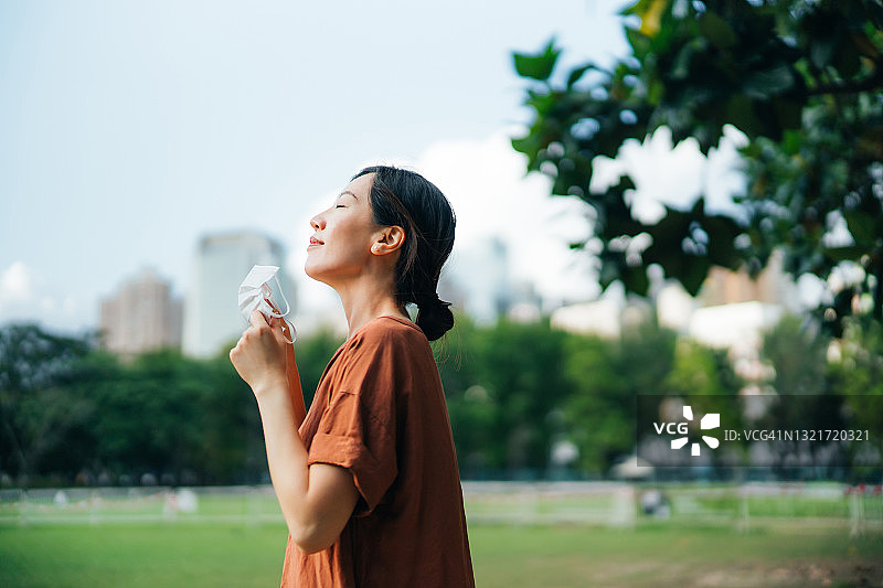 年轻的亚洲女性摘下防护口罩，放松呼吸，享受公园里的新鲜空气，期待疫情结束图片素材