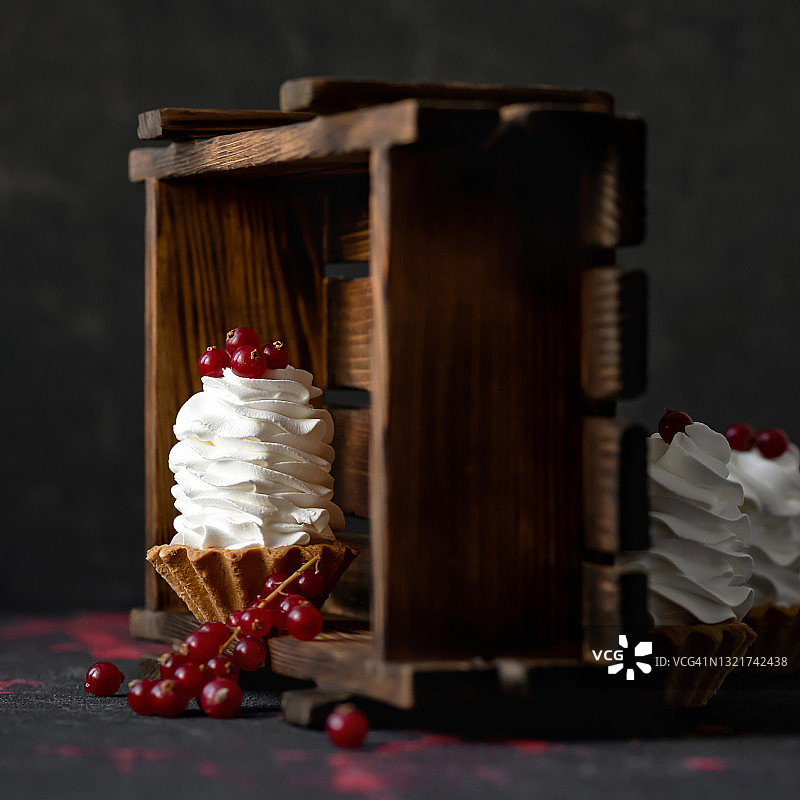 奶油甜点，以红色浆果装饰。烤馅饼或面团篮子，配以透气搅打奶油或慕斯，深色背景，带木箱。迷你蛋糕。复制空间图片素材