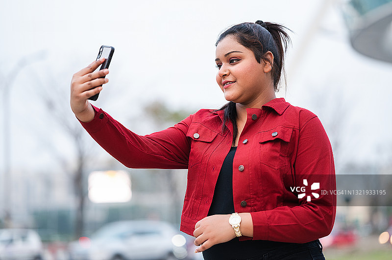 年轻漂亮的微笑女孩正在用智能手机相机自拍。她穿着蓝色牛仔夹克。图片素材