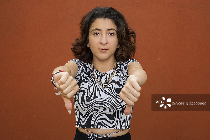 年轻的拉丁裔女人看着镜头，竖起大拇指向下，红色背景图片素材