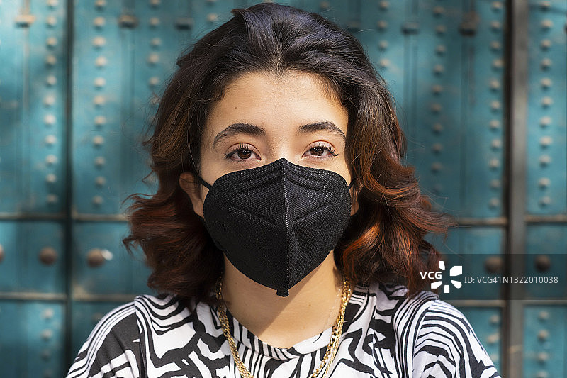 年轻拉丁裔女子戴着口罩看着镜头，绿色背景图片素材