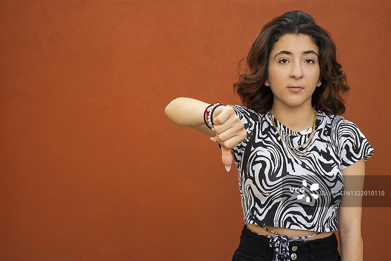 年轻的拉丁裔女人看着镜头，竖起大拇指向下，红色背景图片素材