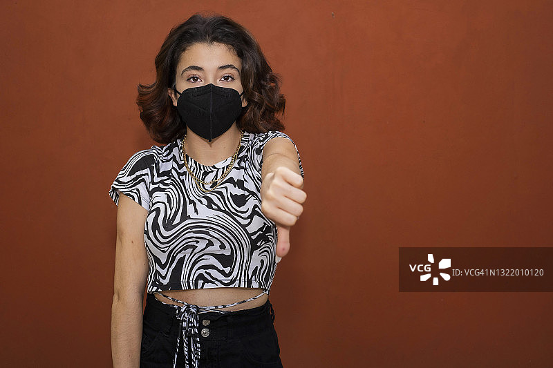 年轻的拉丁裔女子戴着口罩，看着镜头并竖起大拇指向下，红色背景图片素材