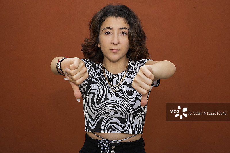 年轻的拉丁裔女人看着镜头，竖起大拇指向下，红色背景图片素材
