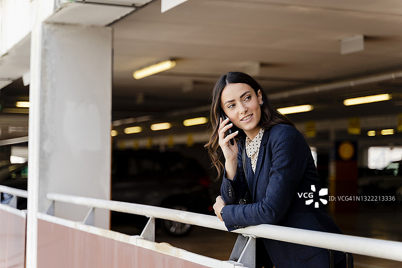 女商人在停车场倚靠在栏杆上，一边看着别处一边用智能手机通话图片素材