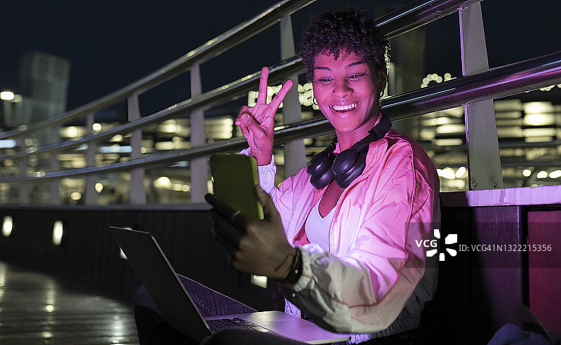 快乐的女人在夜间智能手机视频通话中展示胜利手势图片素材